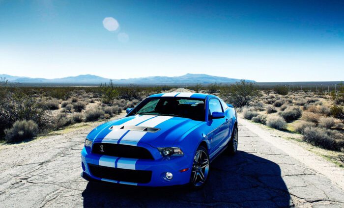 Blue sports car with white racing stripes parked on a desert road under a clear blue sky.