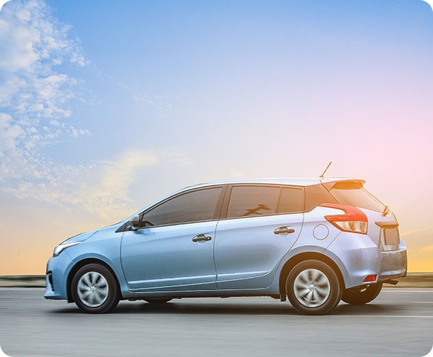 Blue compact car driving on a road under a clear sky at sunset.
